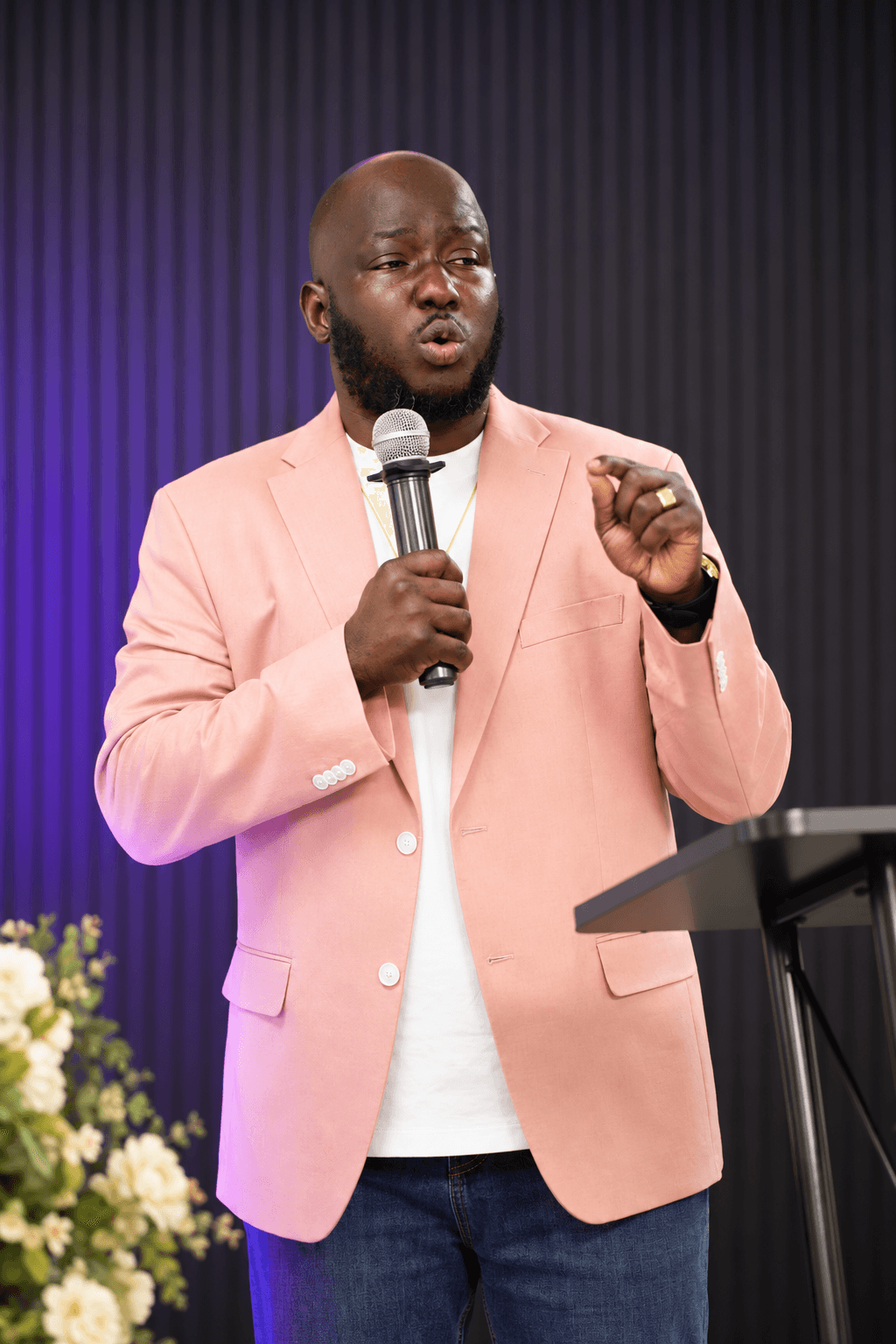 Black man in a pink blazer speaking into a microphone while gesturing at a podium.