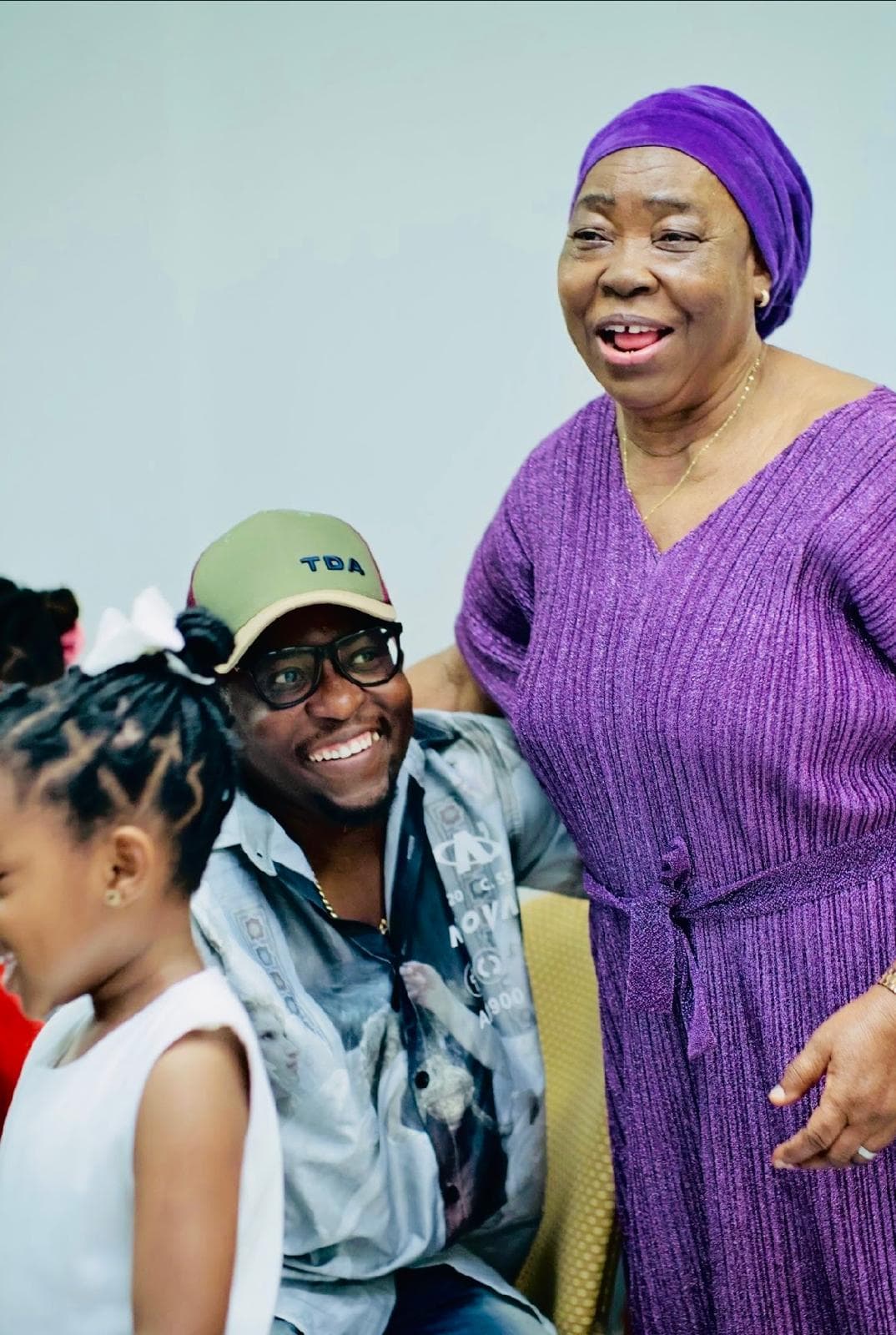 Smiling man in a green cap sits beside an older woman wearing a purple headwrap.
