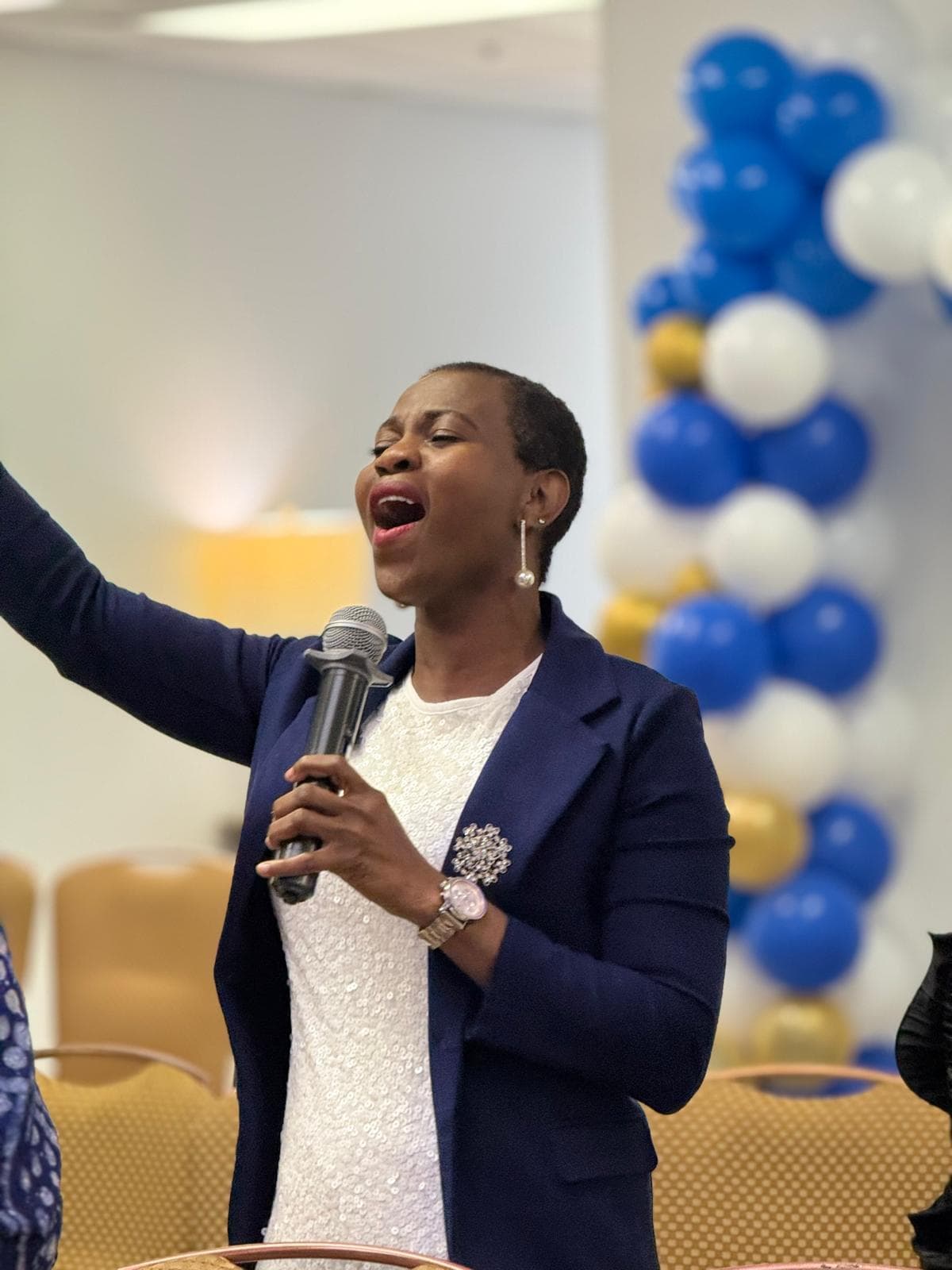 Woman in navy blazer singing into a microphone with arm raised against blue balloon backdrop.