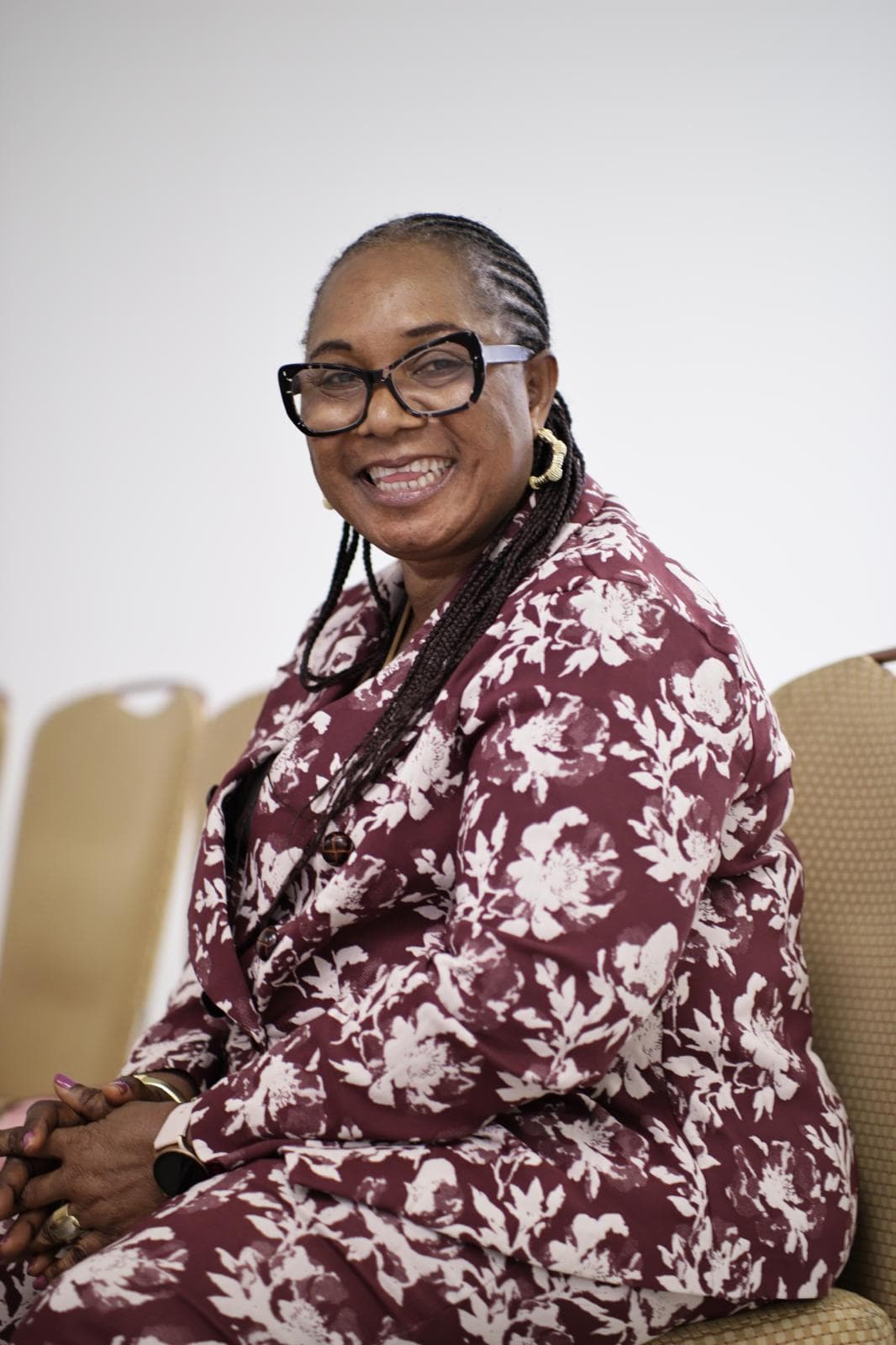 Smiling woman with braided hair and glasses wearing a maroon floral patterned suit, seated.